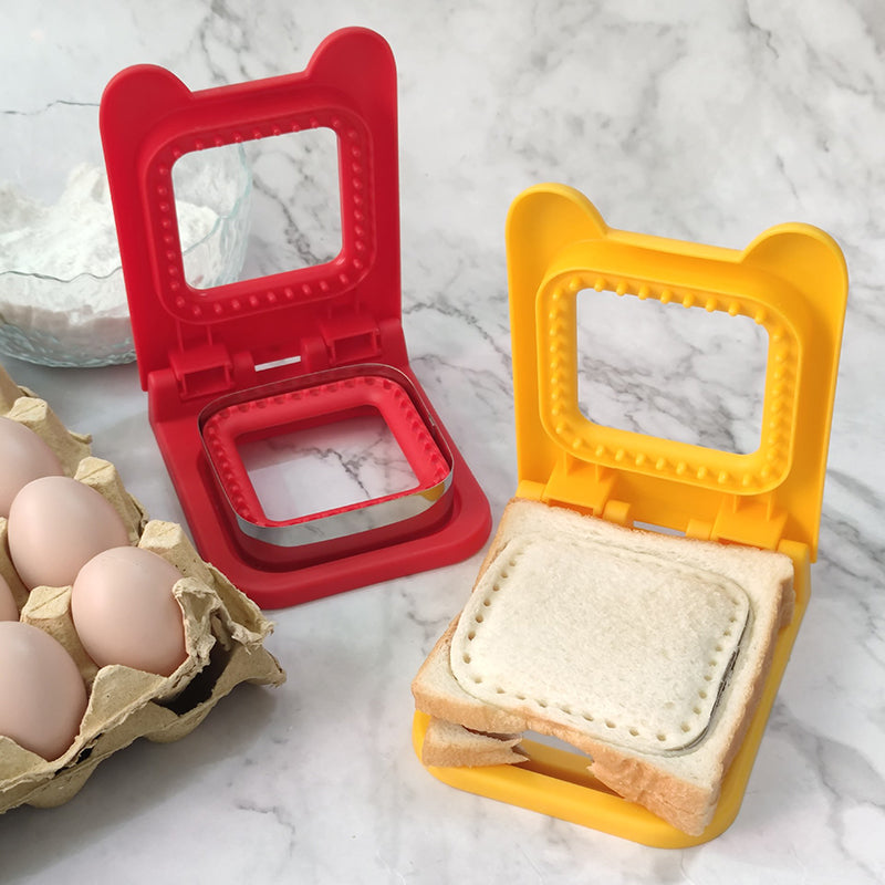 Two sandwich makers, one red and one yellow, on a marble surface with eggs and bread.