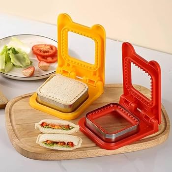 Two sandwich makers, one red and one yellow, on a marble surface with eggs and bread.