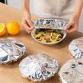 Person unwrapping a bowl of food covered with tinfoil on a wooden table with oranges in the background.