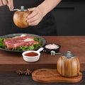 Wooden pepper grinder being used over a plate of raw meat on a wooden table.