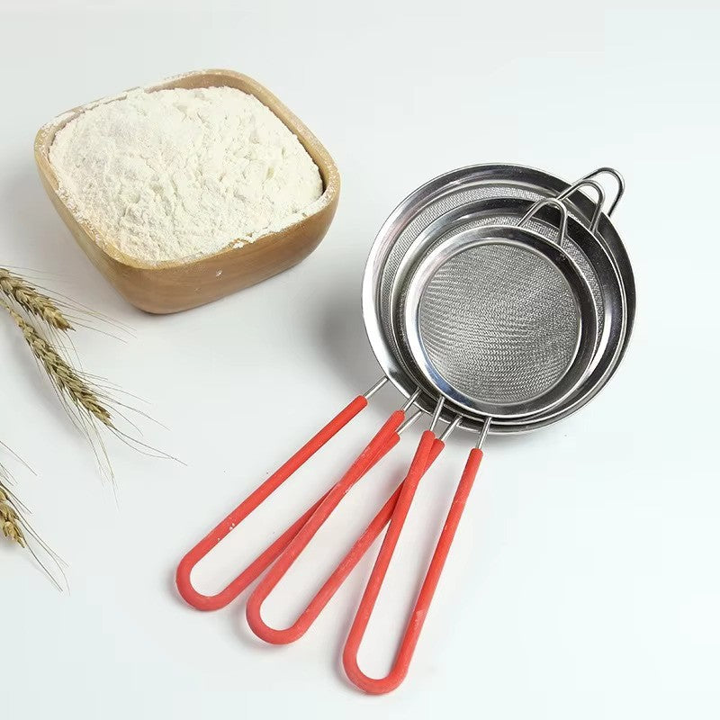 Three metal sieves with red handles next to a bowl of flour on a white background