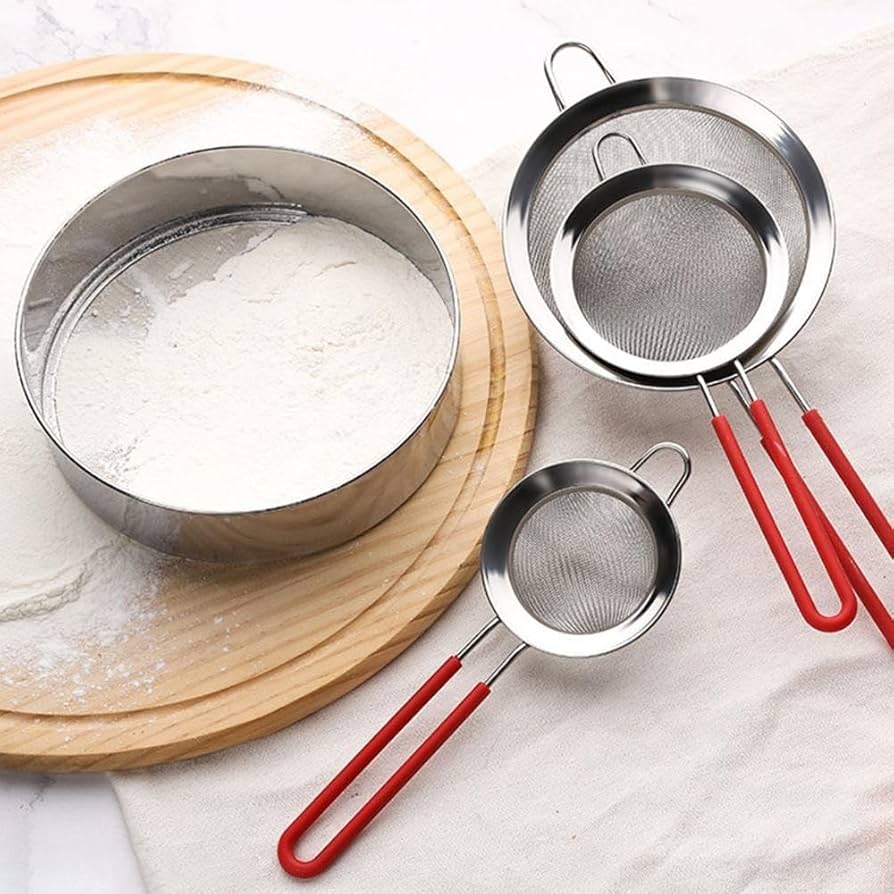 Three metal sifters with red handles on a white surface, one sifting flour.