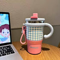Pink and checkered insulated mug with handle on a wooden desk next to a laptop.